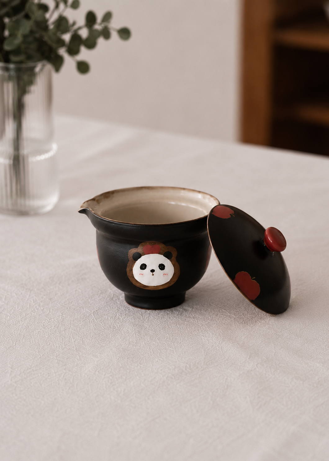 A black ceramic tea bowl with a matching lid, decorated with a cute panda face and red accents, set on a light beige tablecloth with a blurred background.