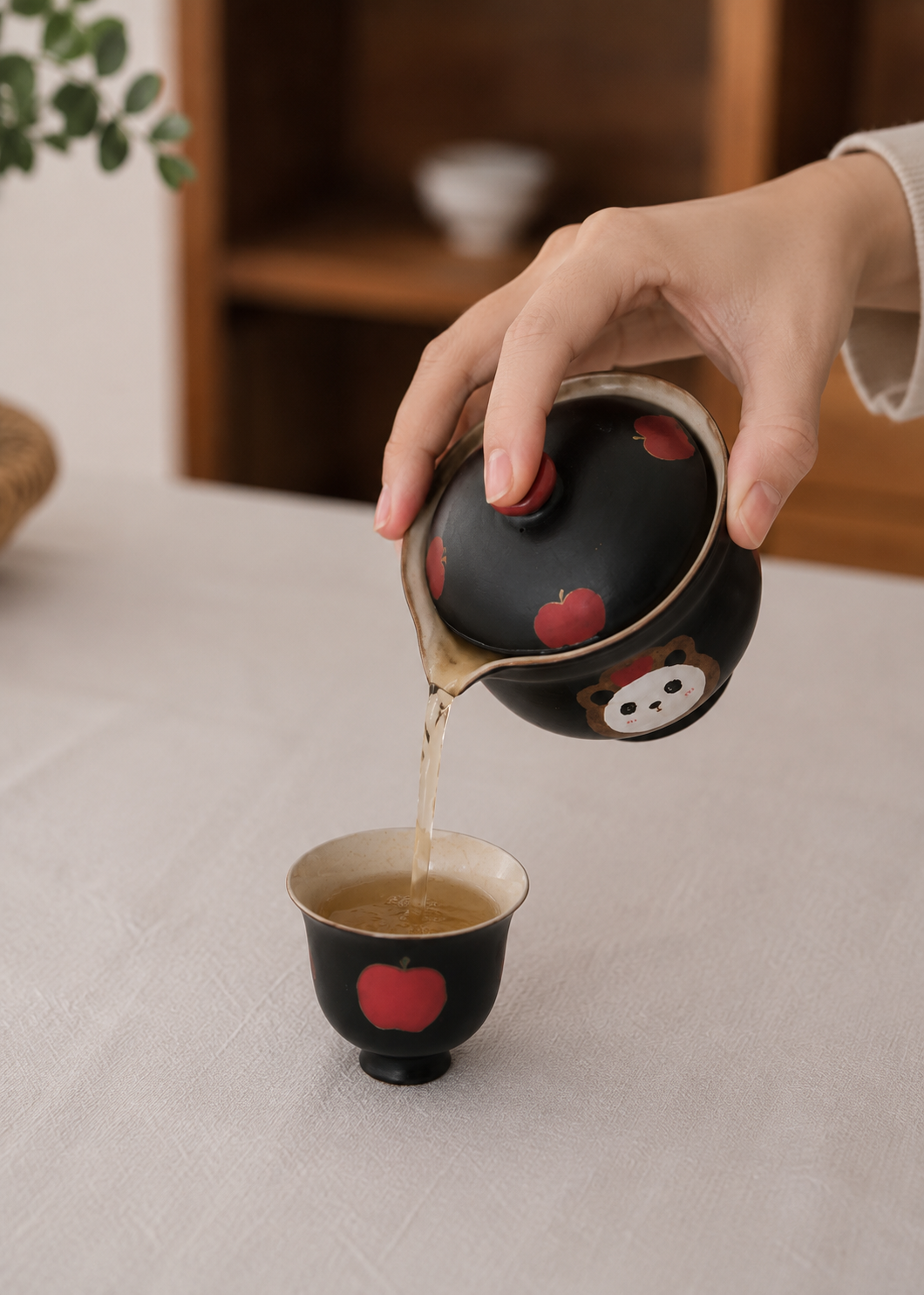 A hand pouring tea from a black ceramic teapot decorated with red apples and a cartoon face into a matching small tea cup on a light tablecloth.