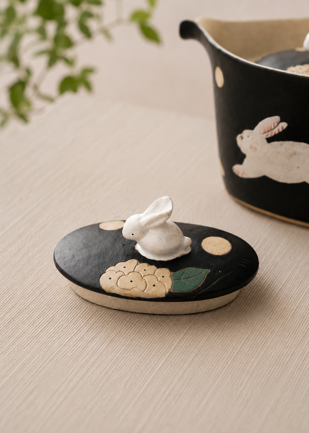 A close-up of a black ceramic lid with a raised white rabbit figure and cream-colored decorative details, set on a light beige textured surface with a matching black ceramic container blurred in the background.