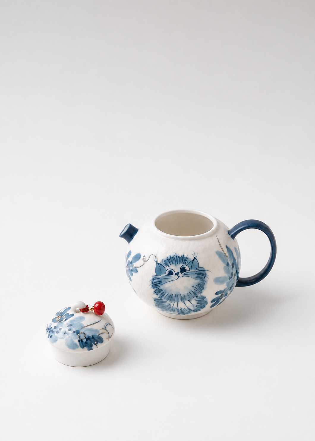 A white ceramic teapot with a blue cat illustration and blue handle, shown with its matching lid on a light gray background.