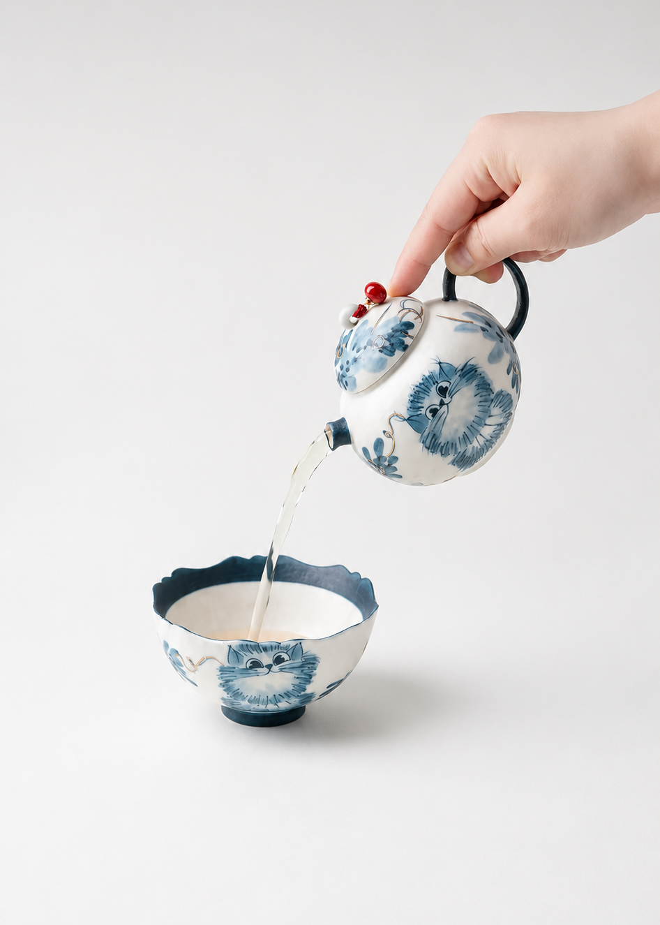 A hand with red nail polish holds a small blue-and-white floral teapot pouring liquid into a matching blue-and-white bowl, set against a clean light gray background.