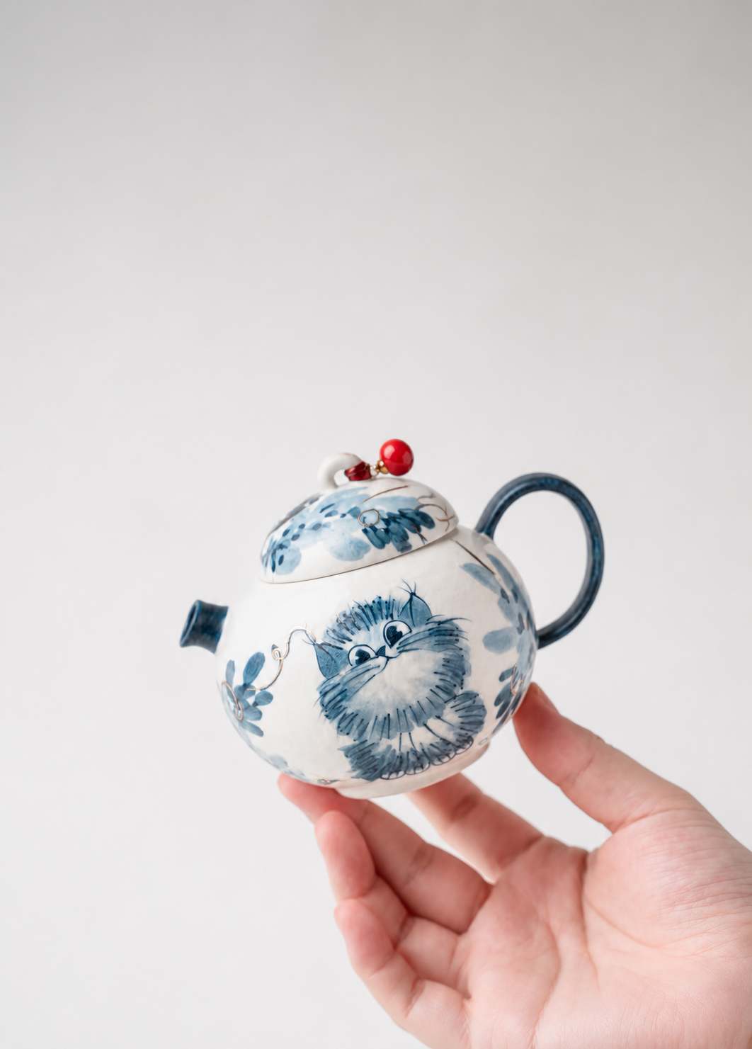 A hand holding a small white ceramic teapot decorated with blue cat illustrations and a red berry accent on the lid, against a plain light gray background.