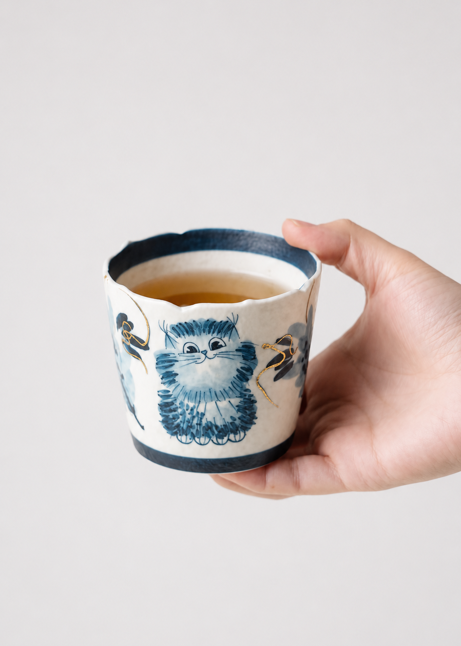 A hand holding a small ceramic cup with a blue and white owl illustration and a dark rim, filled with tea, against a plain light gray background.