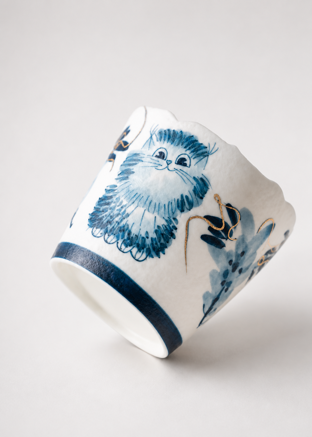 A white ceramic cup with a hand-painted blue and gold illustration of a fluffy cat, shown on a light gray background.