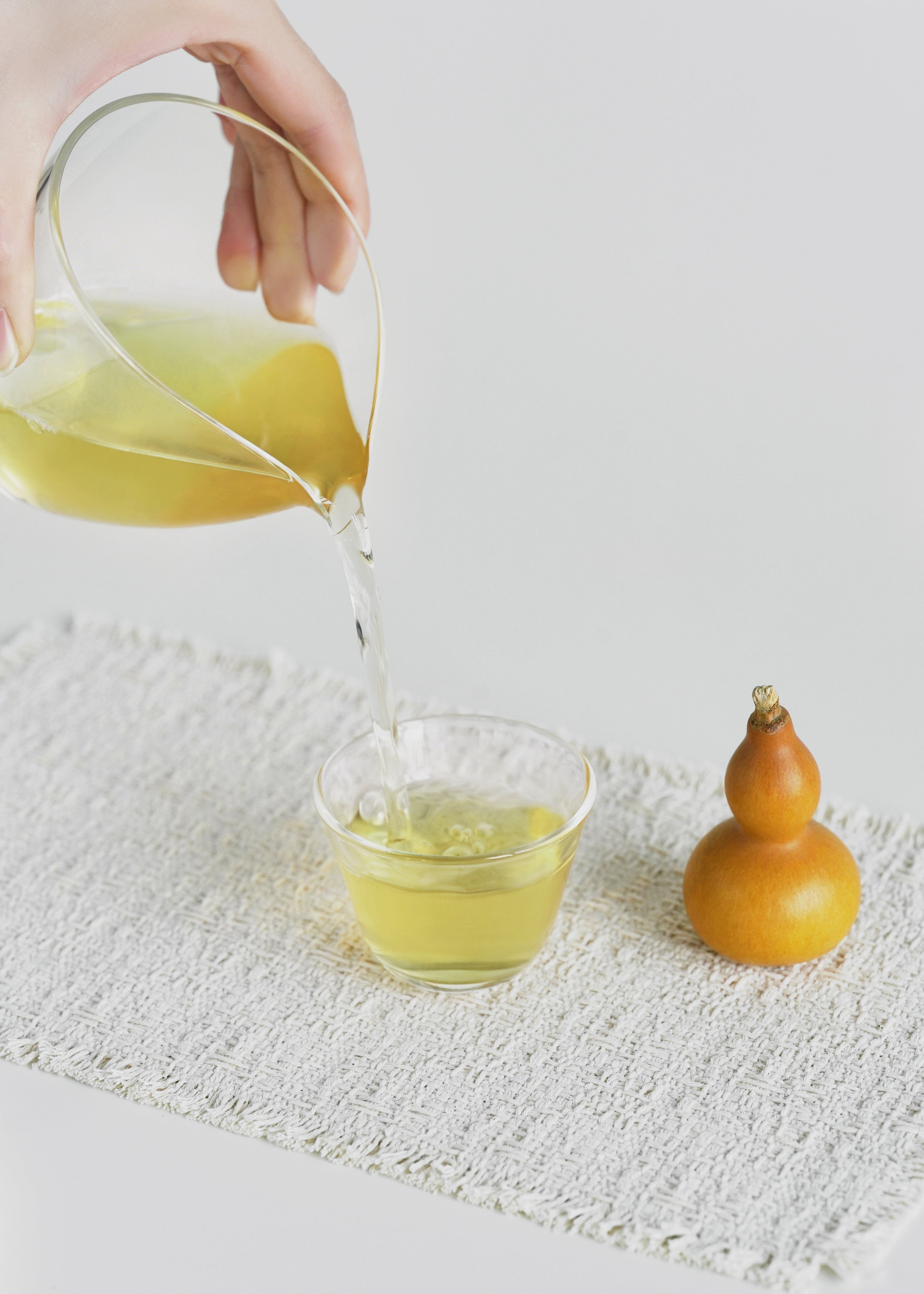 Hand pouring a yellow liquid from a pitcher into a glass on a textured white surface with a small gourd. Jasmine green tea