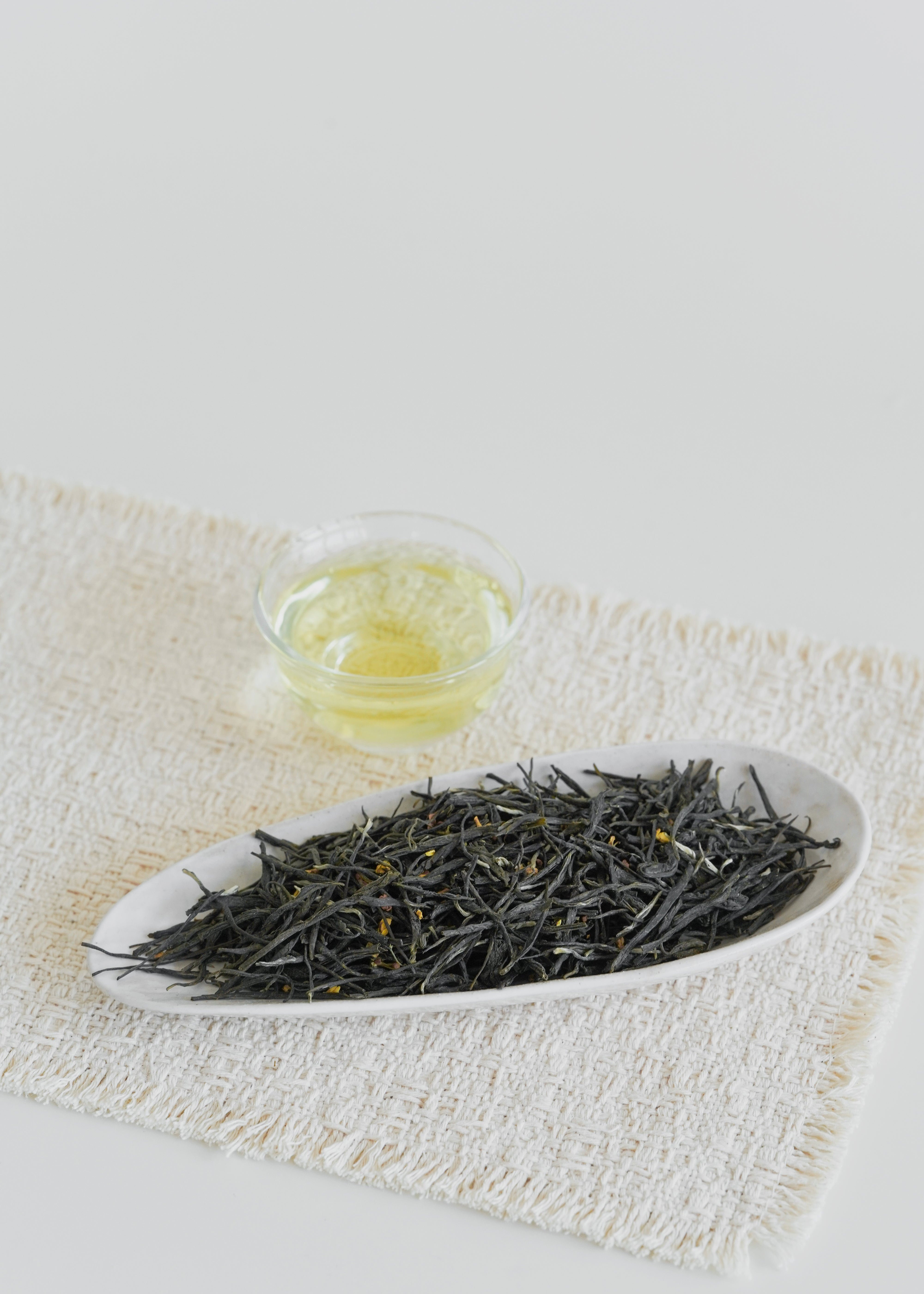 Tea leaves in a white dish with a small glass of tea liquid on a light background. Osmanthus Green Tea