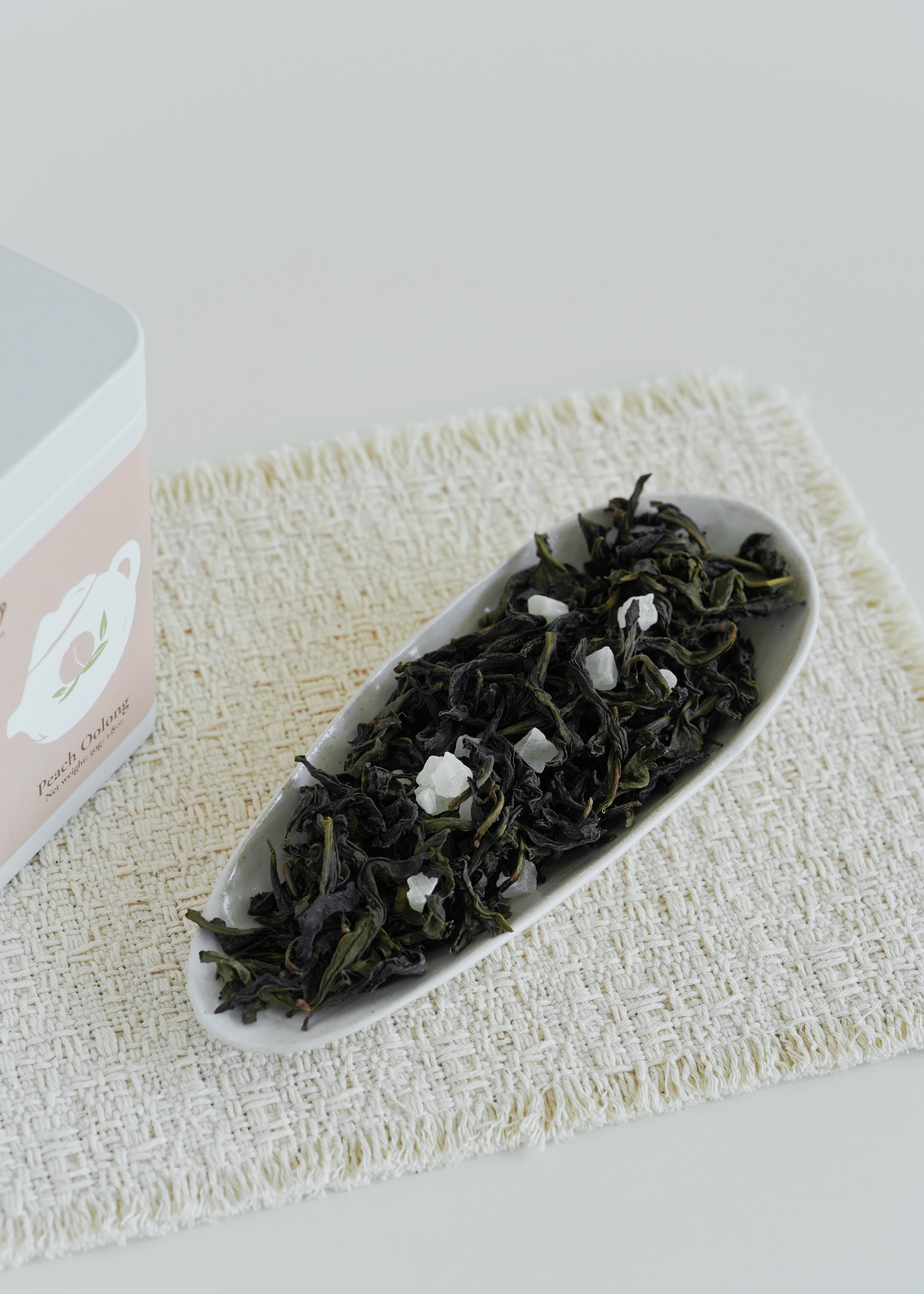 Tea leaves in a white dish on a textured mat with a pink box in the background. Peach Oolong