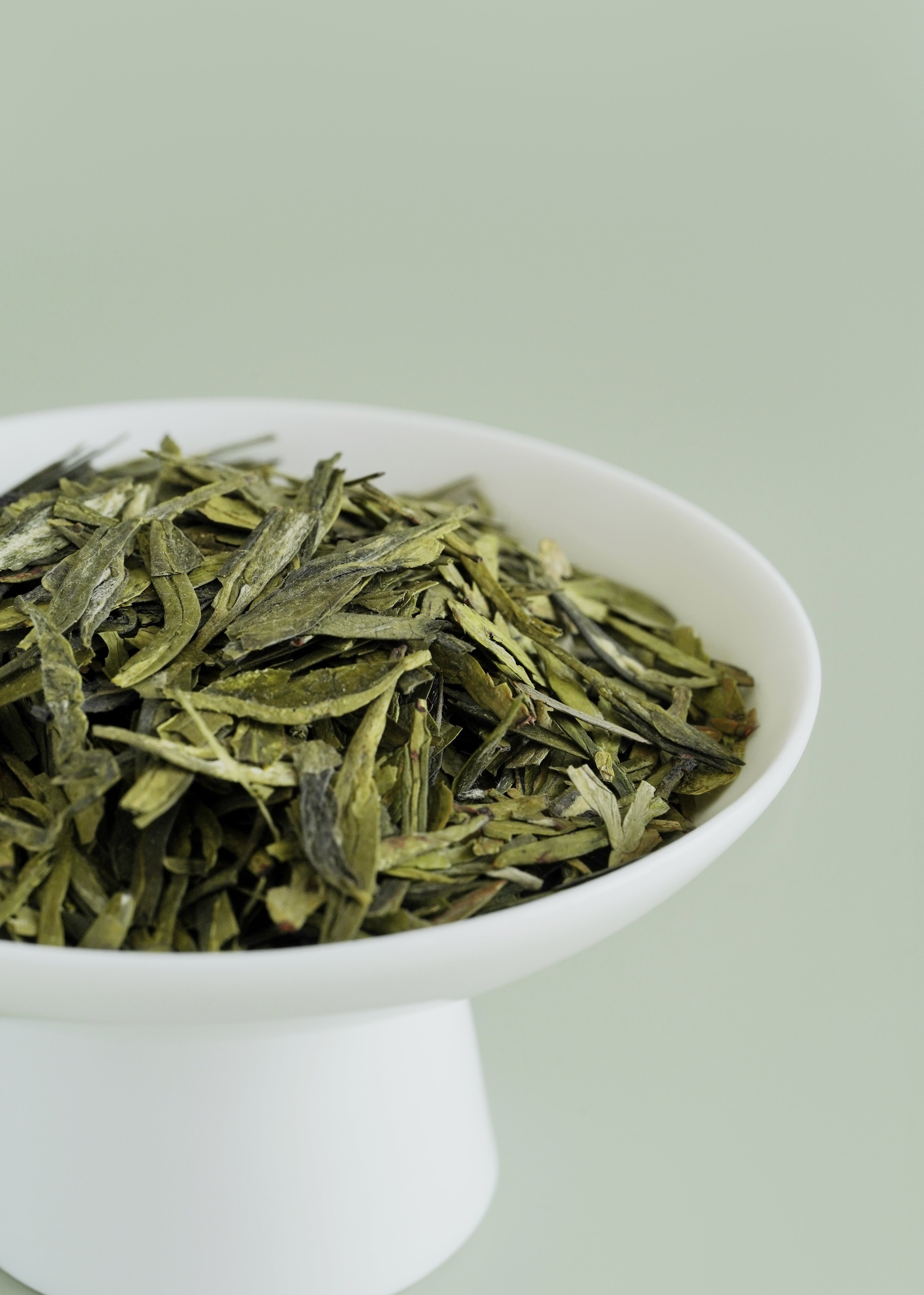 Green tea leaves in a white bowl on a light green background. Dragon well longjing