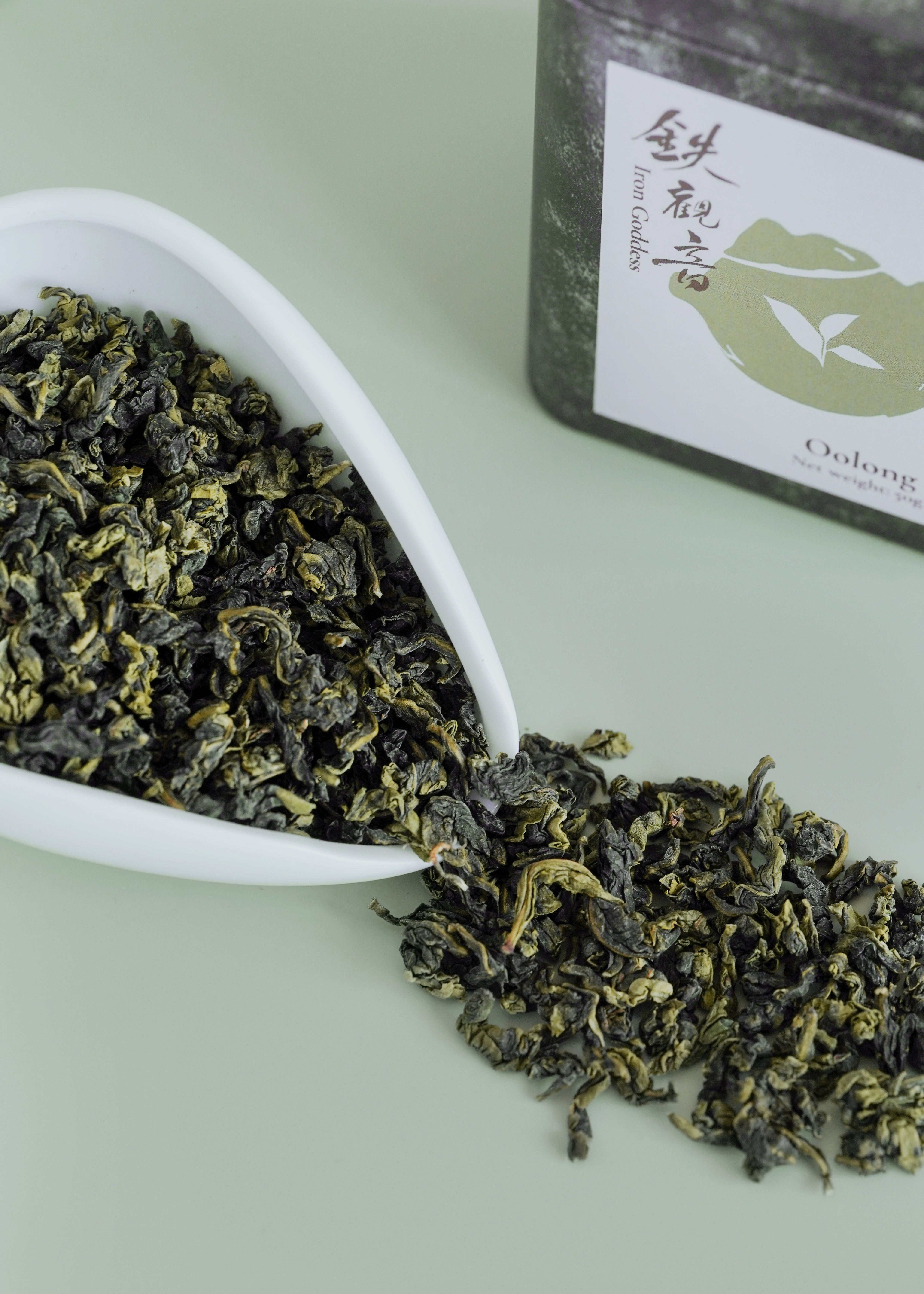 Oolong tea leaves spilling from a white scoop onto a light green surface with a package of Oolong tea in the background. Iron goddess
