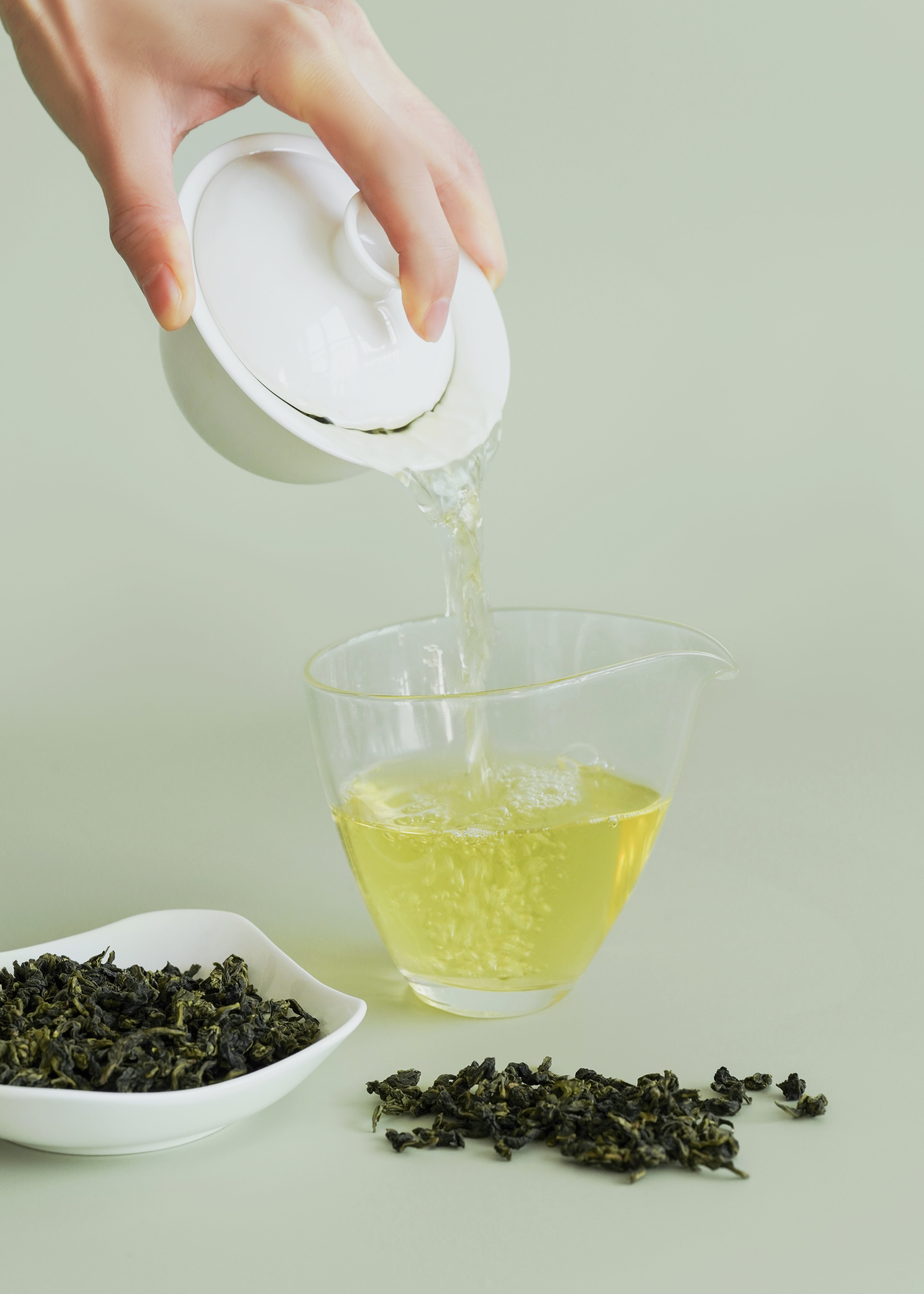 Hand pouring green tea into a glass with tea leaves and a tea bag on a light green background. Iron goddess