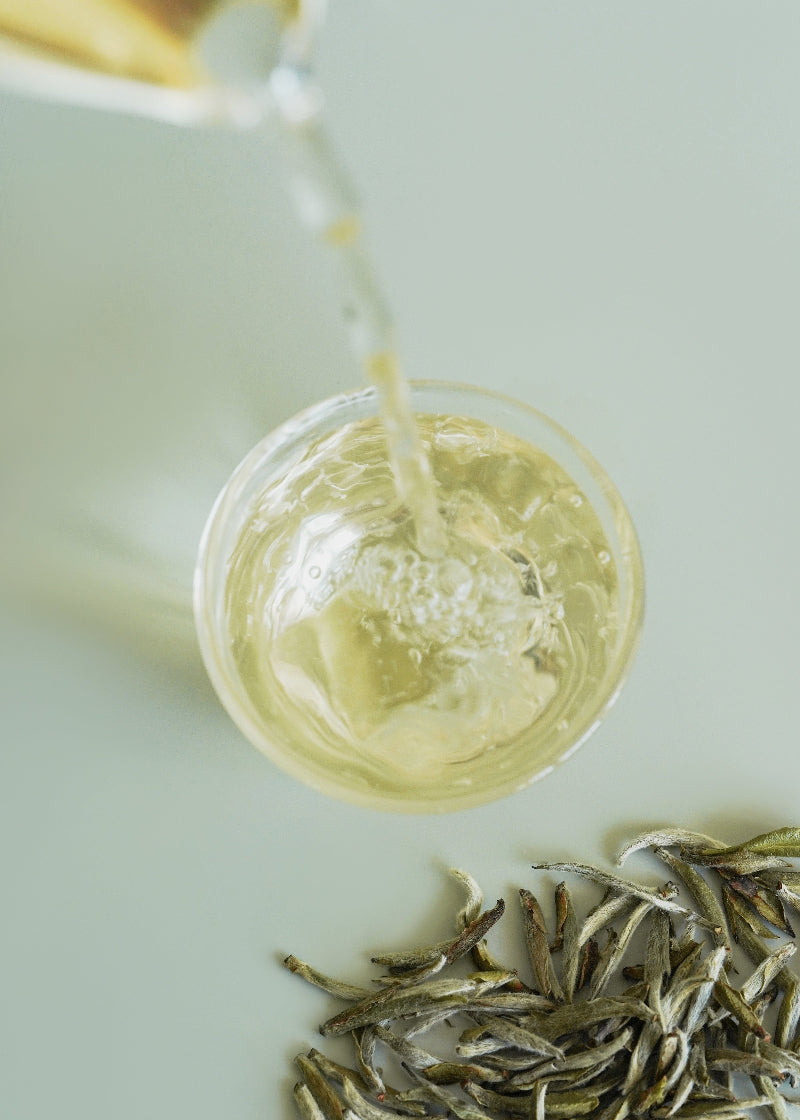 Tea being poured into a cup with tea leaves on a light surface. SIlver needle