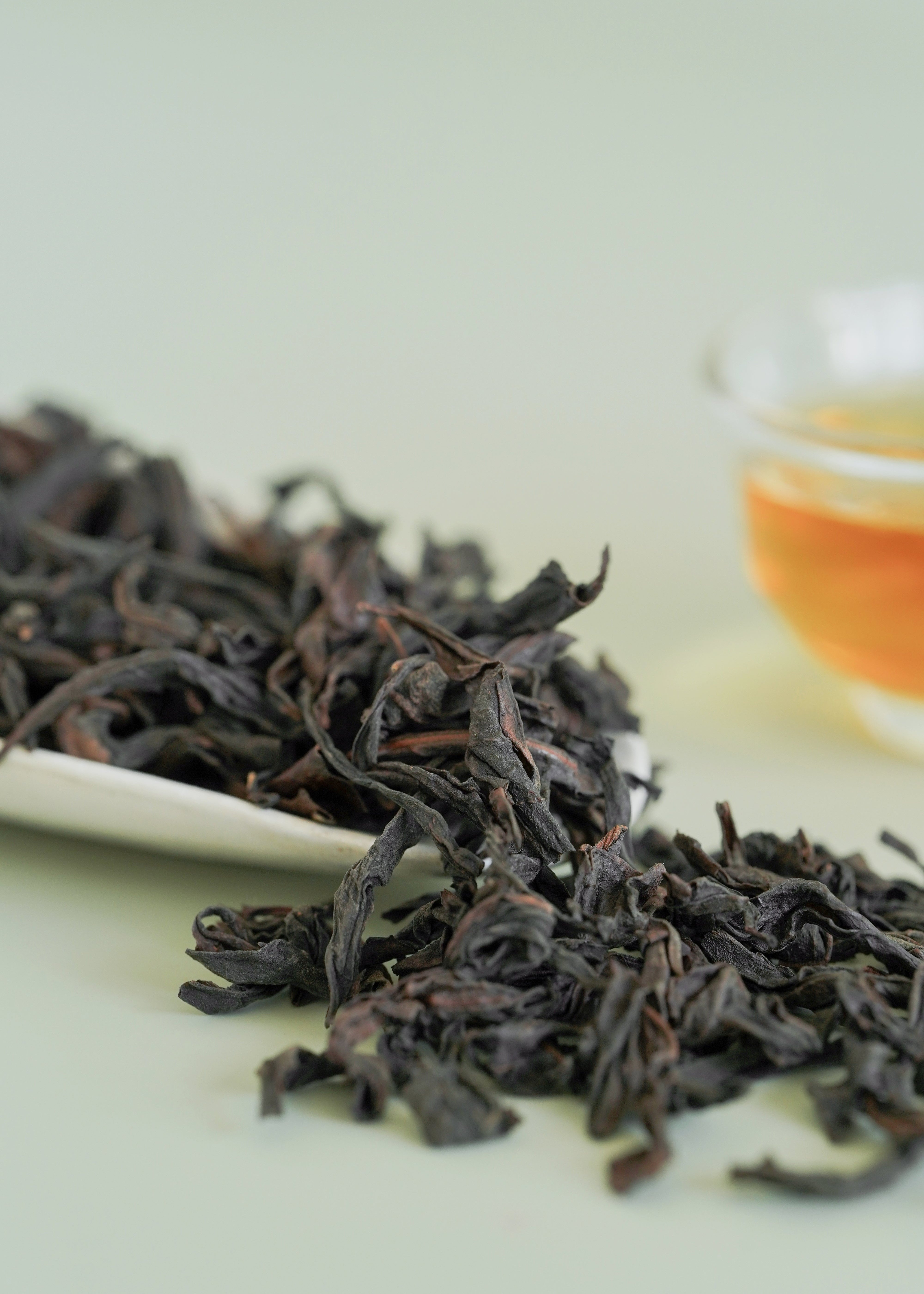 Dried black tea leaves on a white plate with a blurred glass of tea in the background. Big red robe, da hong pao