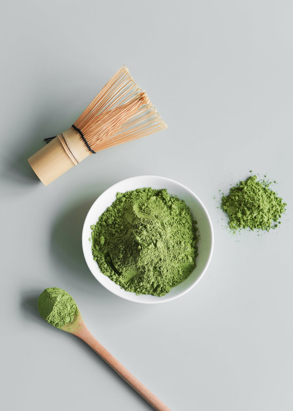 Matcha green tea powder in a white bowl with a whisk, spoon, and scattered matcha on a light gray background.
