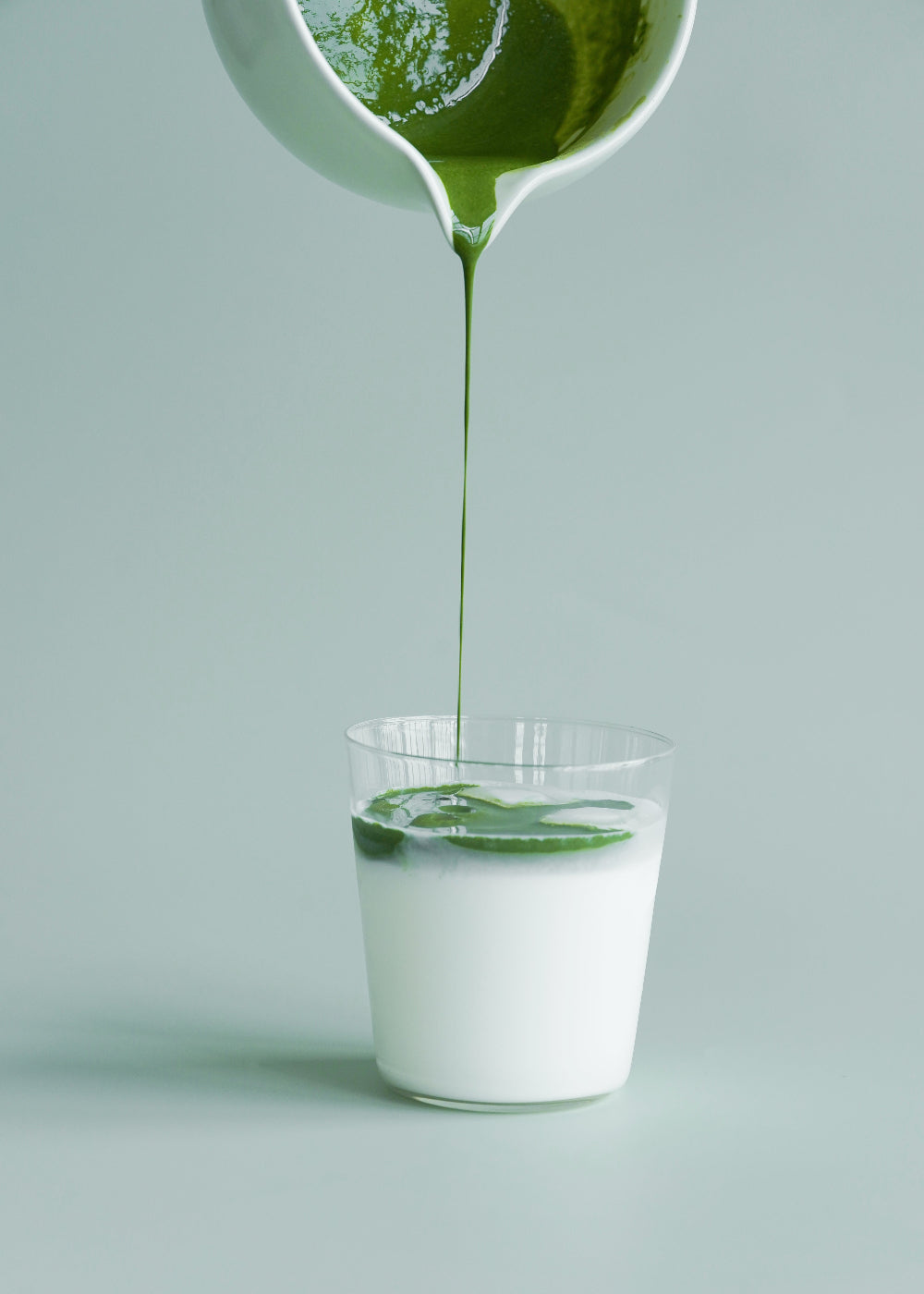 Green liquid being poured from a pitcher into a clear glass on a light gray background