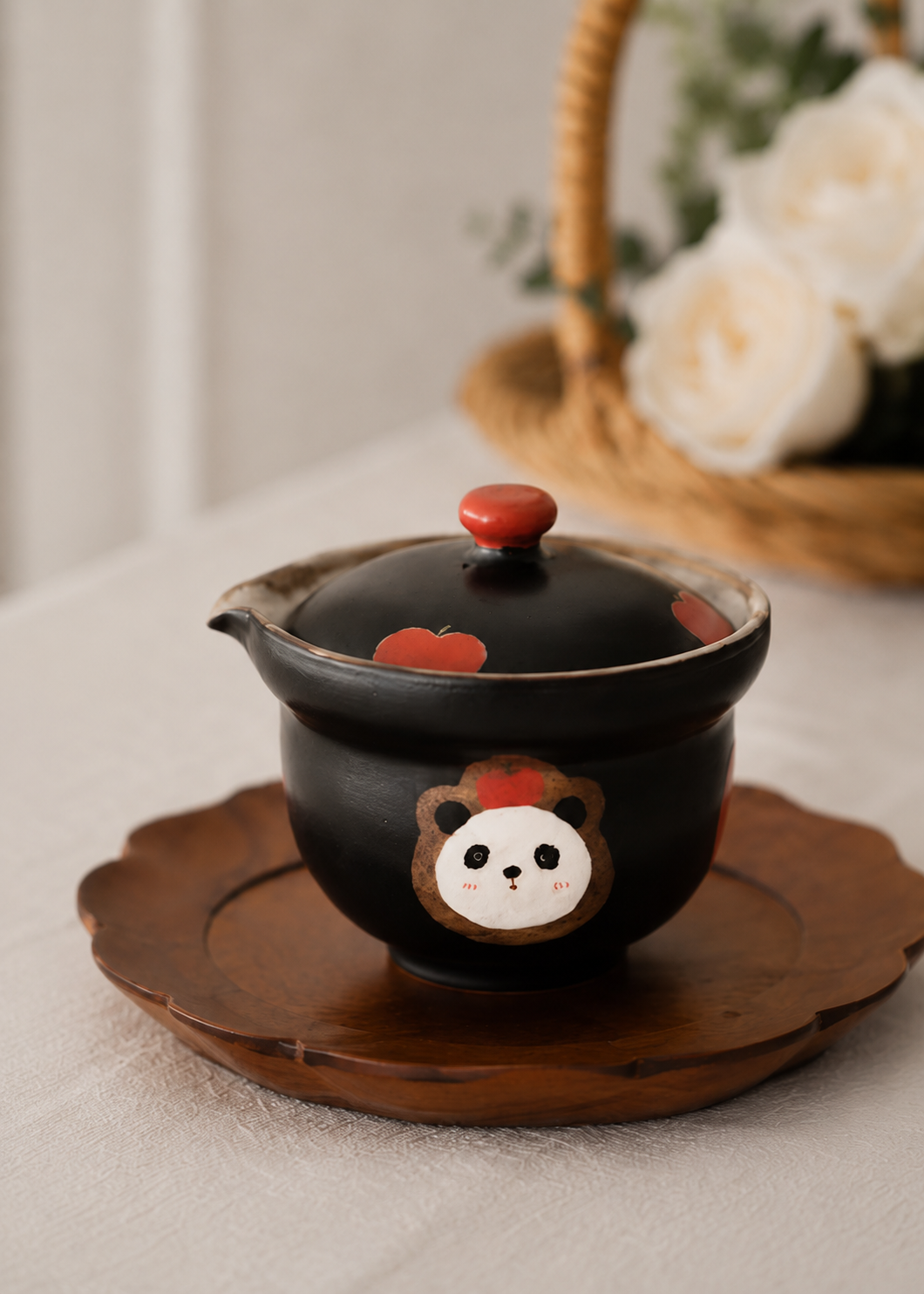 A black ceramic teapot with a matching lid, decorated with a cute bear face and red apple motifs, resting on a scalloped wooden tray. The background is softly blurred with a woven basket and white flowers.