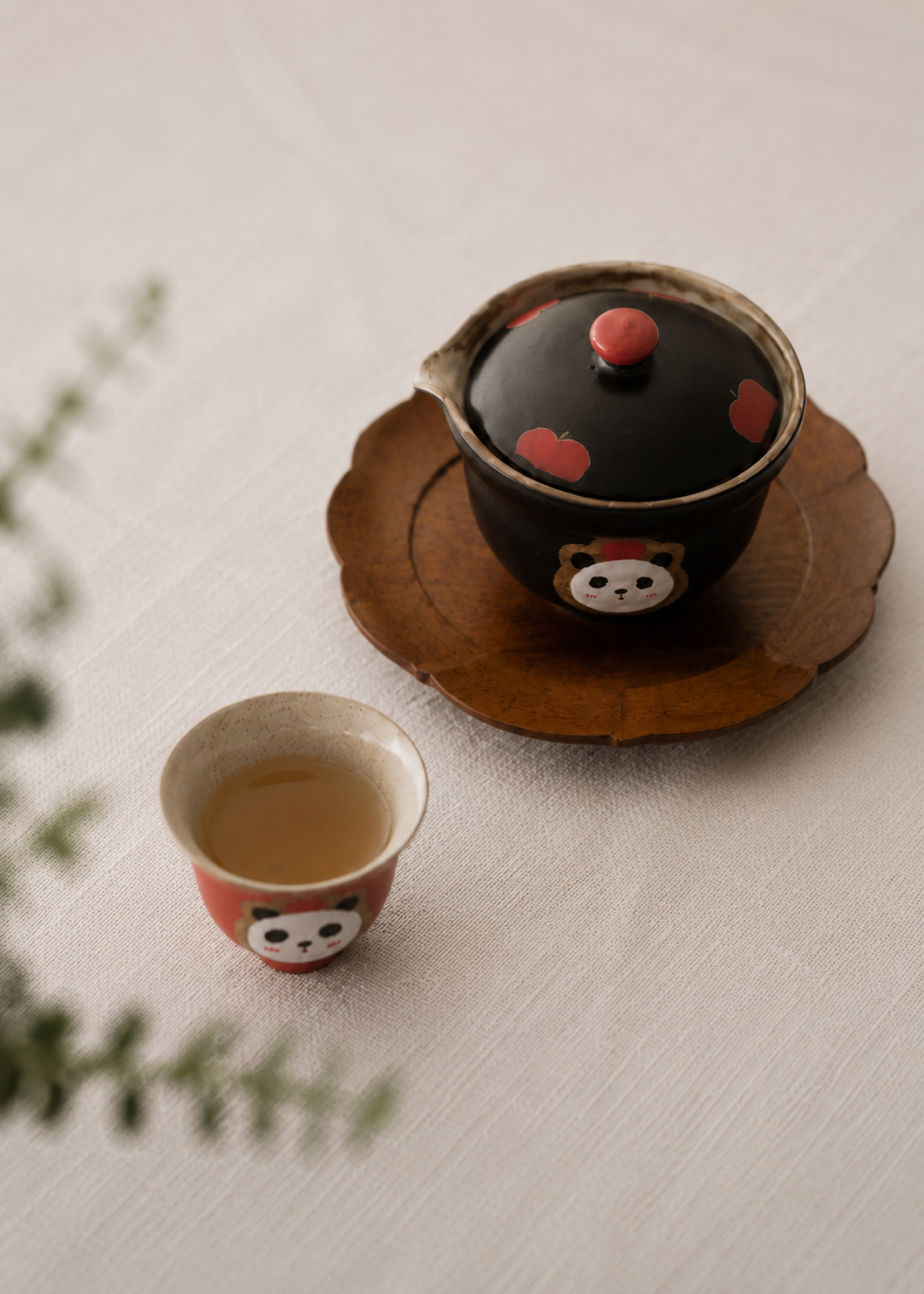 A ceramic tea set with a black lid decorated with red spots and a bear face, shown on a wooden tray next to a matching small teacup filled with tea on a light fabric surface.