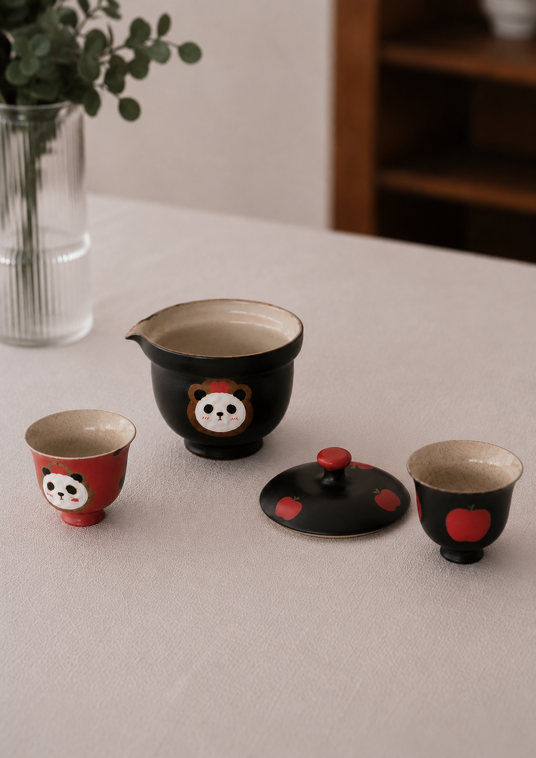 A ceramic tea set on a light beige tablecloth, featuring a black gaiwan with a panda face, a matching black lid with red spots, a small red cup with a panda face, and a small black cup with a red apple face. A glass vase with green leaves is blurred in the background.