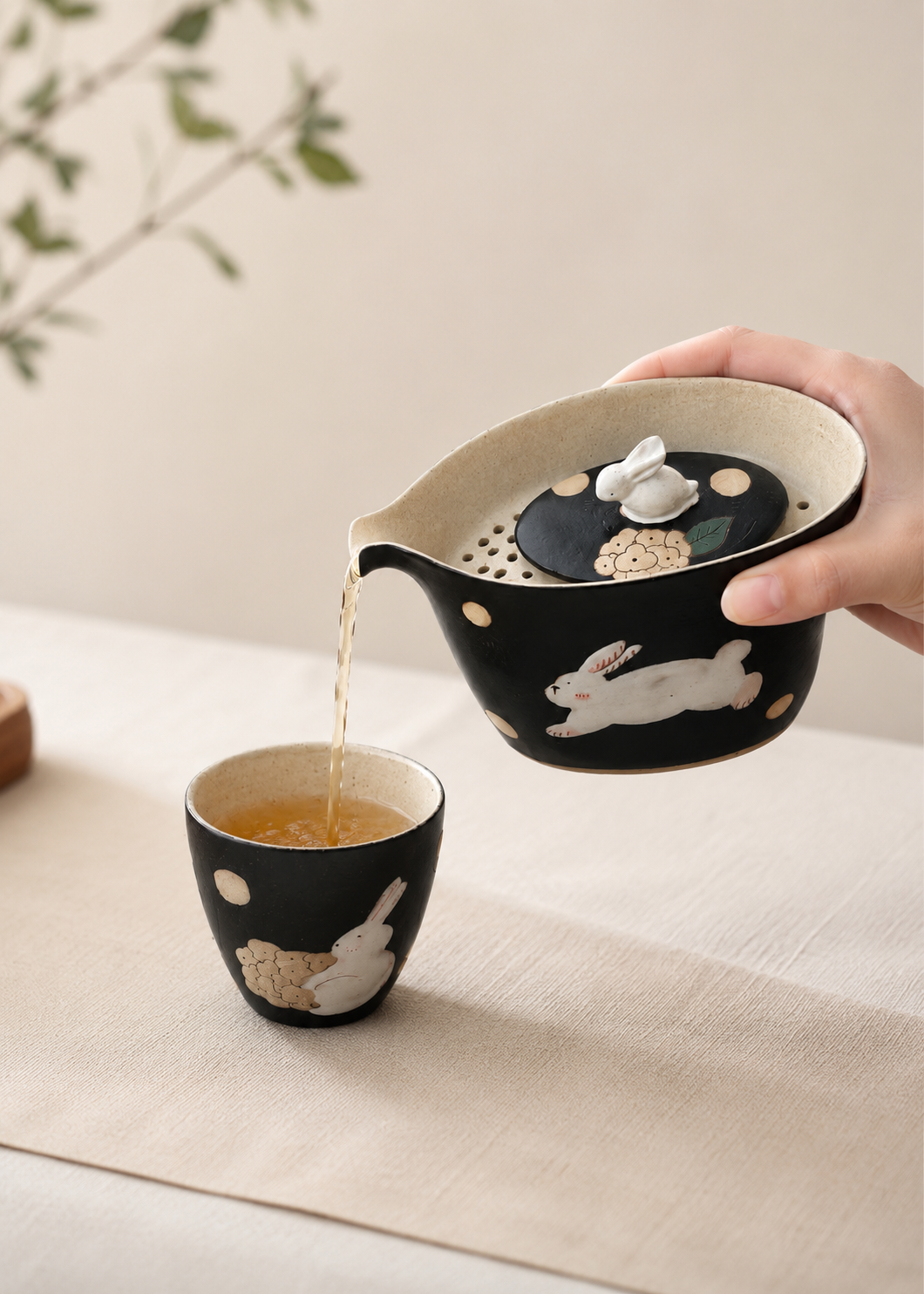 A hand holds a black ceramic teapot with white rabbit illustrations and gold polka dots, pouring tea into a matching small cup on a beige table runner.