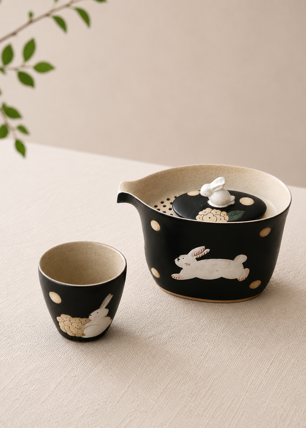 A black ceramic tea set with white rabbit illustrations and beige speckled details, including a teapot with a lid and a matching small cup, styled on a light beige surface with a soft neutral background.