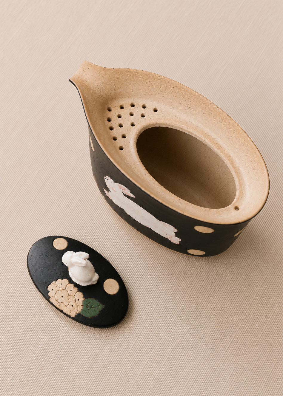 A ceramic teapot with a matching lid, featuring a black exterior decorated with white rabbit illustrations and beige polka dots, and a natural light wood-toned interior. The teapot is shown on a textured beige background next to its lid.