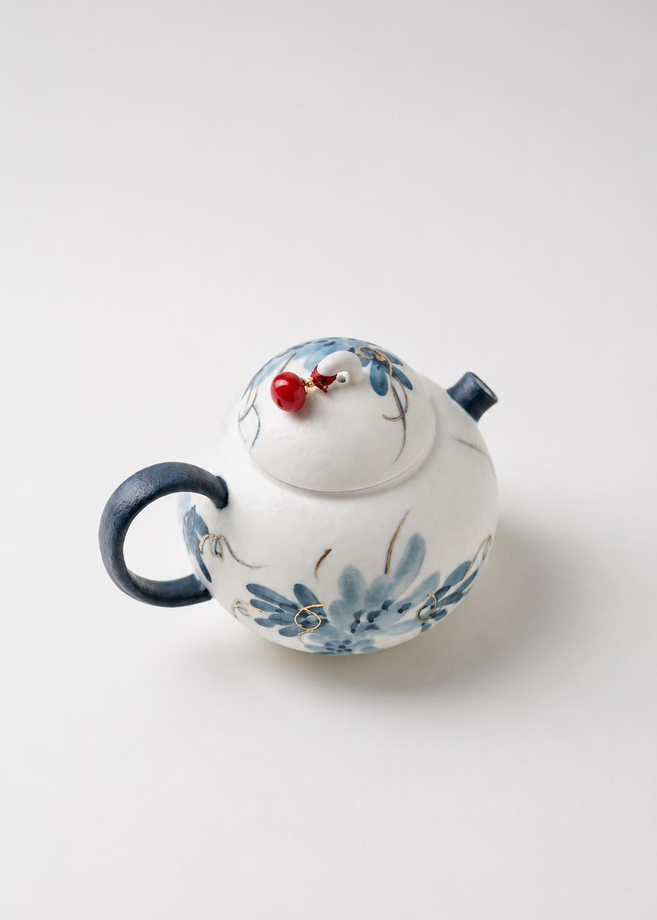 A small white ceramic teapot with blue floral hand-painted patterns, a dark blue handle, and a lid topped with a small red decorative accent, shown on a light gray background.