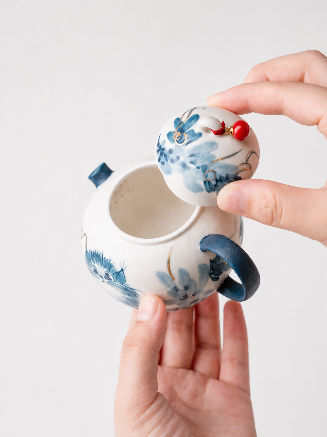 A small white ceramic teapot with blue floral hand-painted details, held by two hands against a light gray background. The right hand is lifting the lid, which features a small red decorative bead accent.