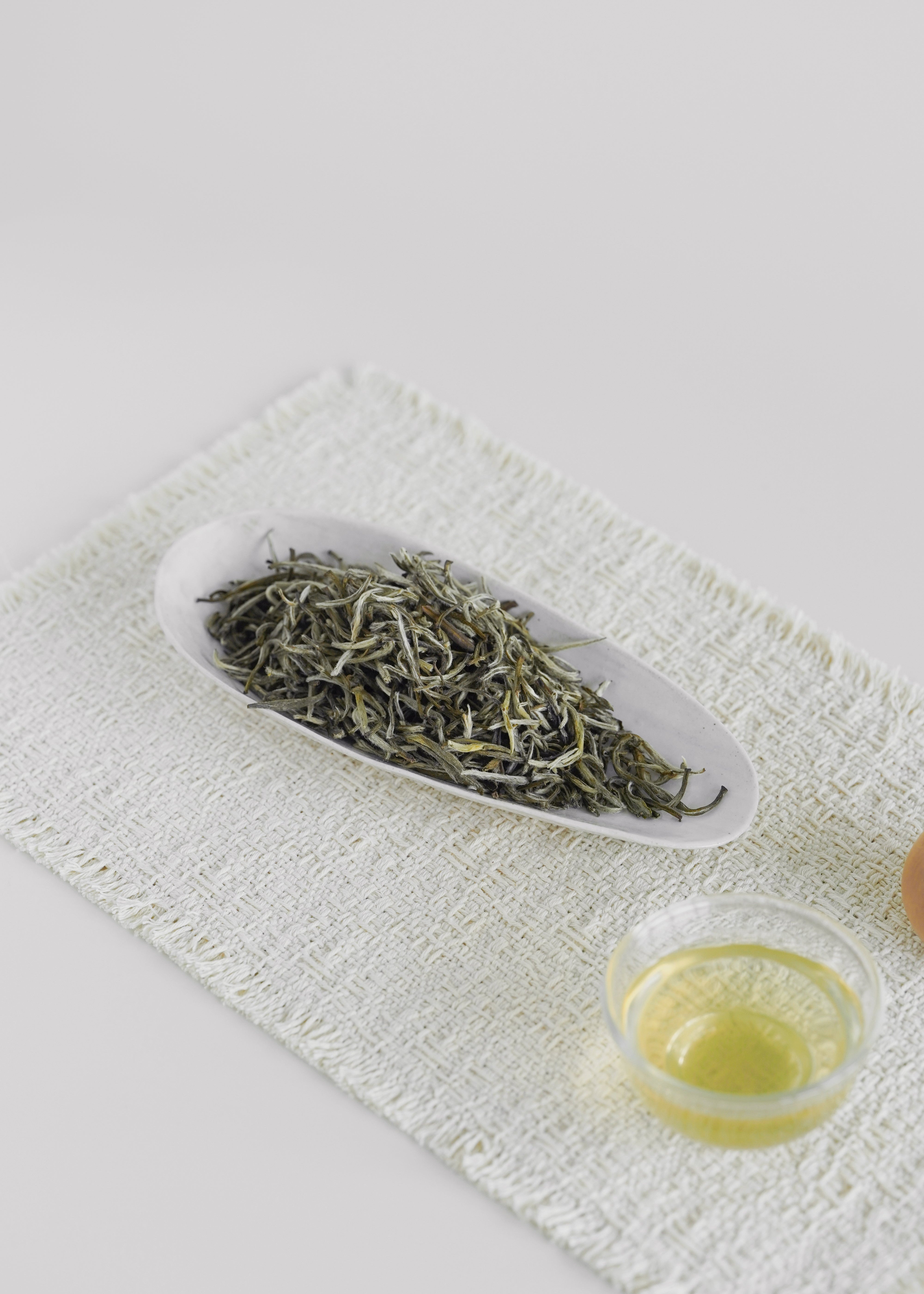Dry green tea leaves on a white dish with a small glass of green liquid on a textured white surface. Jasmine green tea