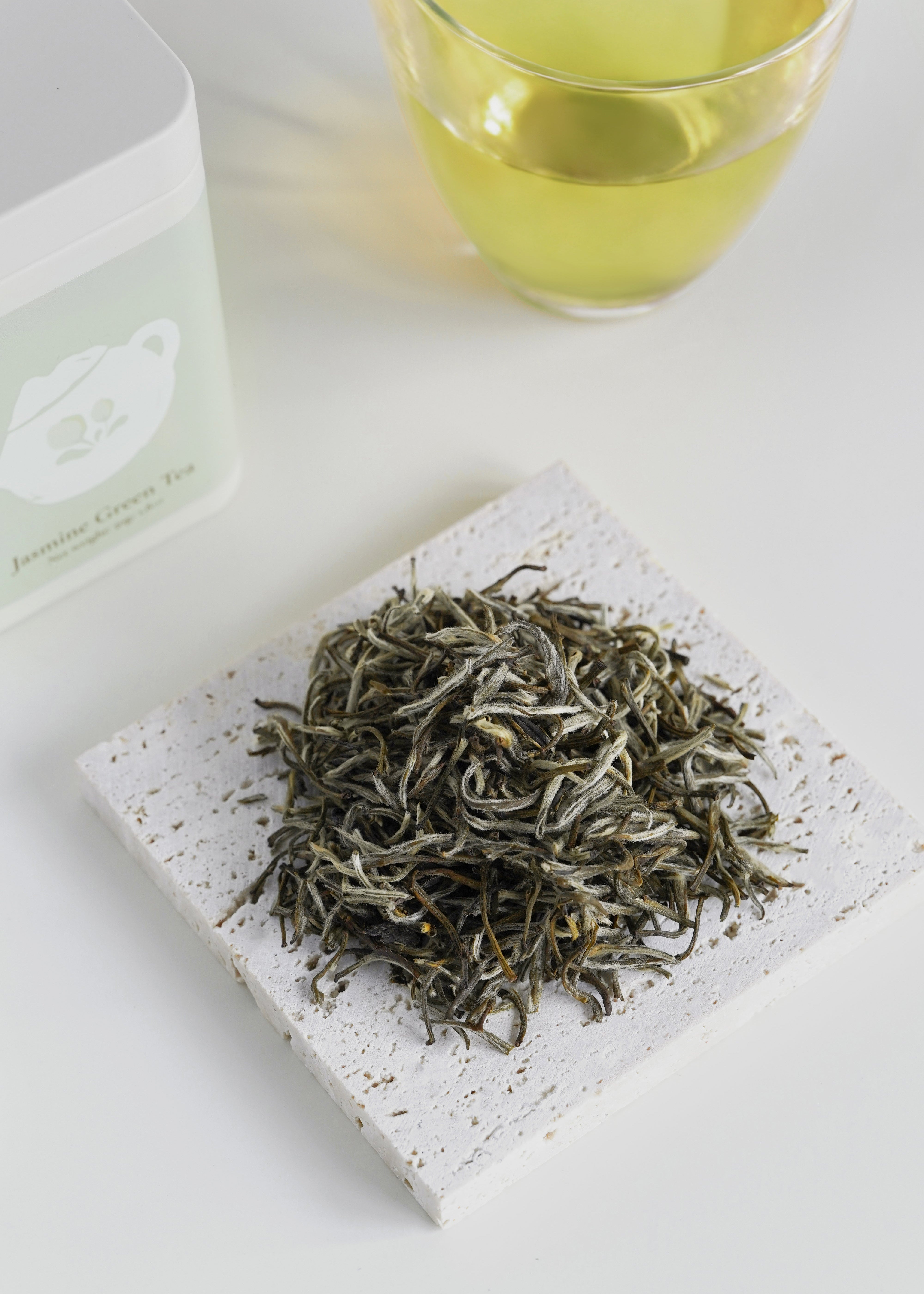 Dry green tea leaves on a stone surface with a glass of tea in the background. Jasmine green tea