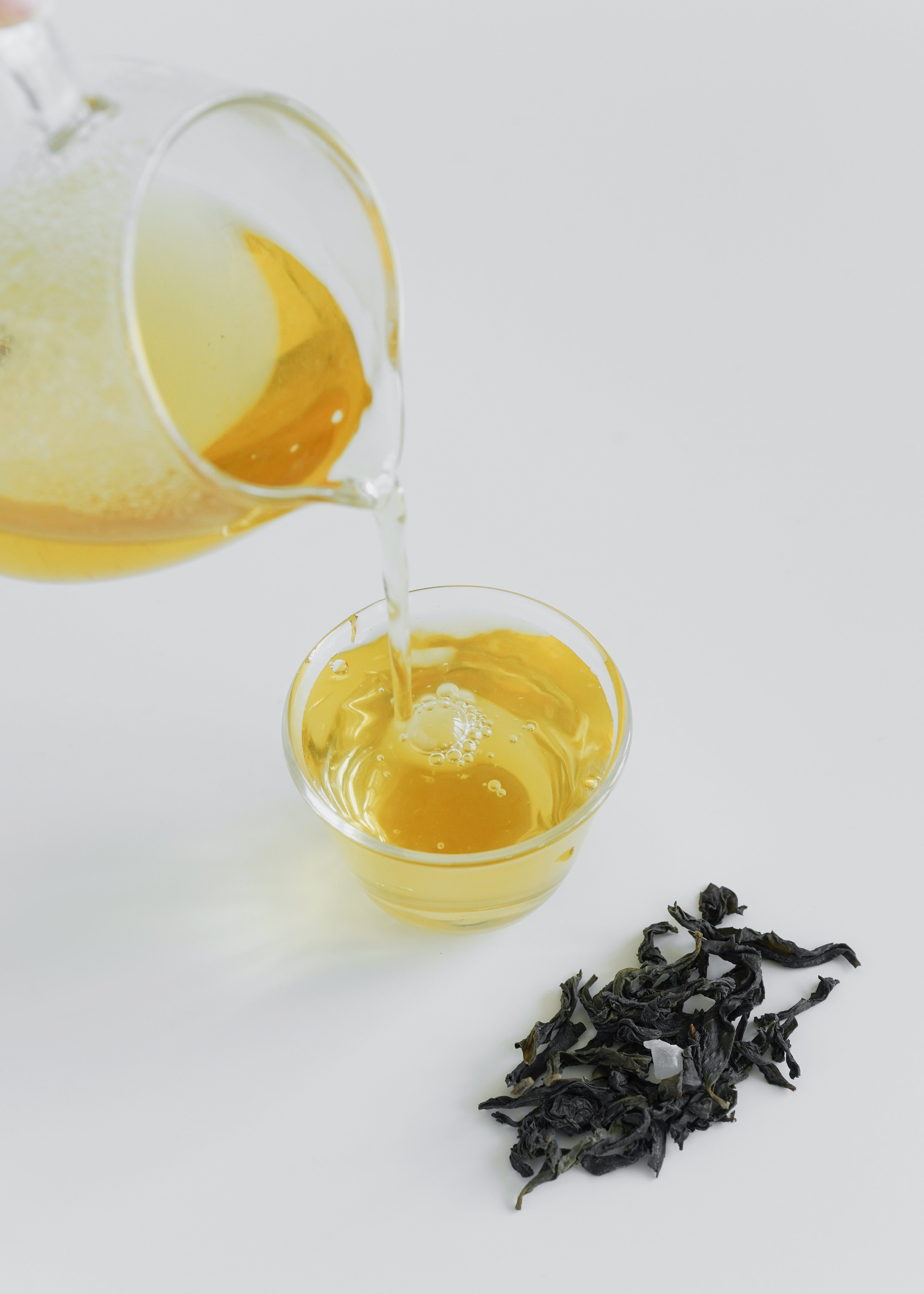 Tea being poured from a glass teapot into a small glass cup with tea leaves on a white background.  Peach Oolong