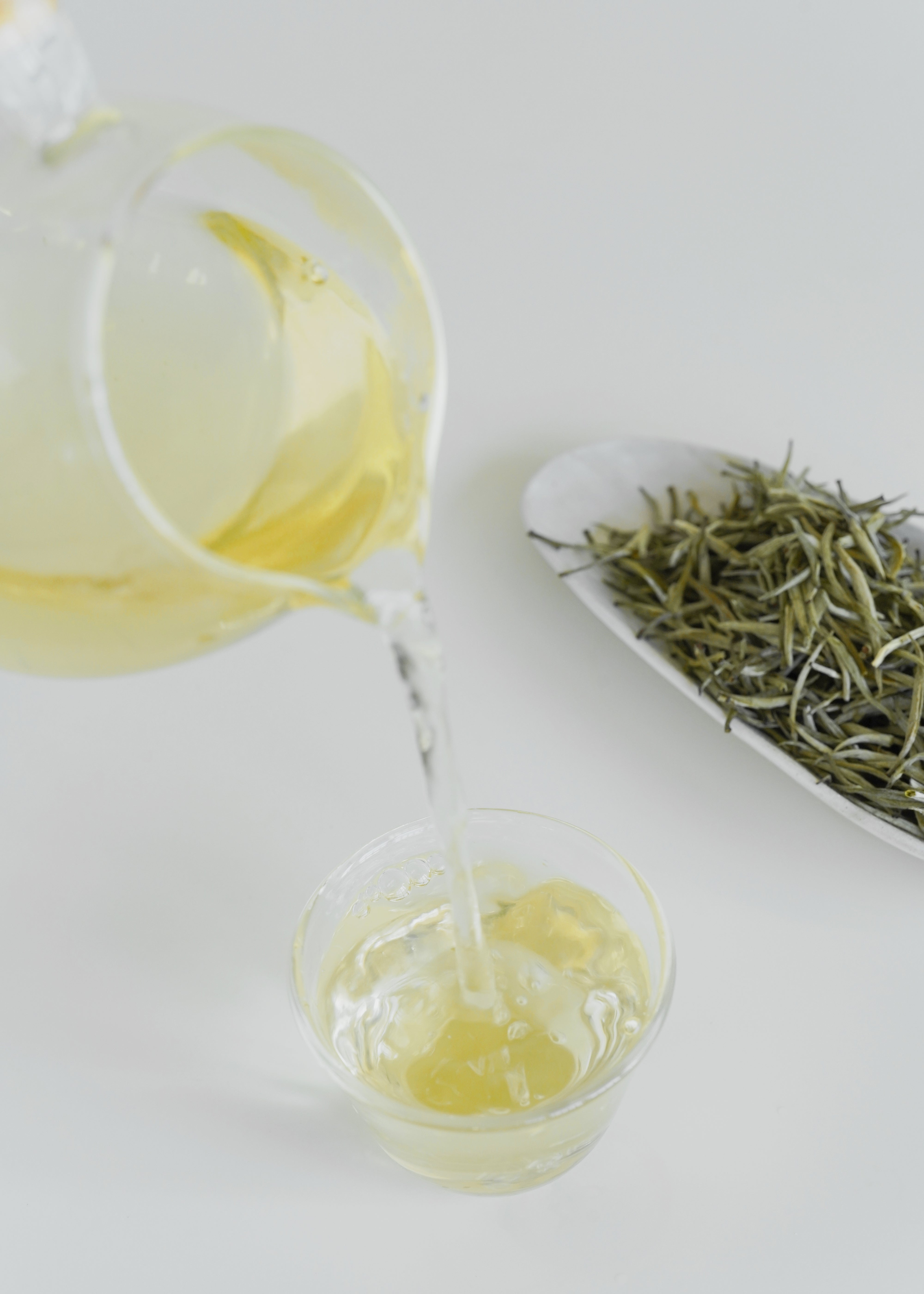 Tea being poured from a teapot into a glass cup with tea leaves on a white surface. Orchid yellow tea