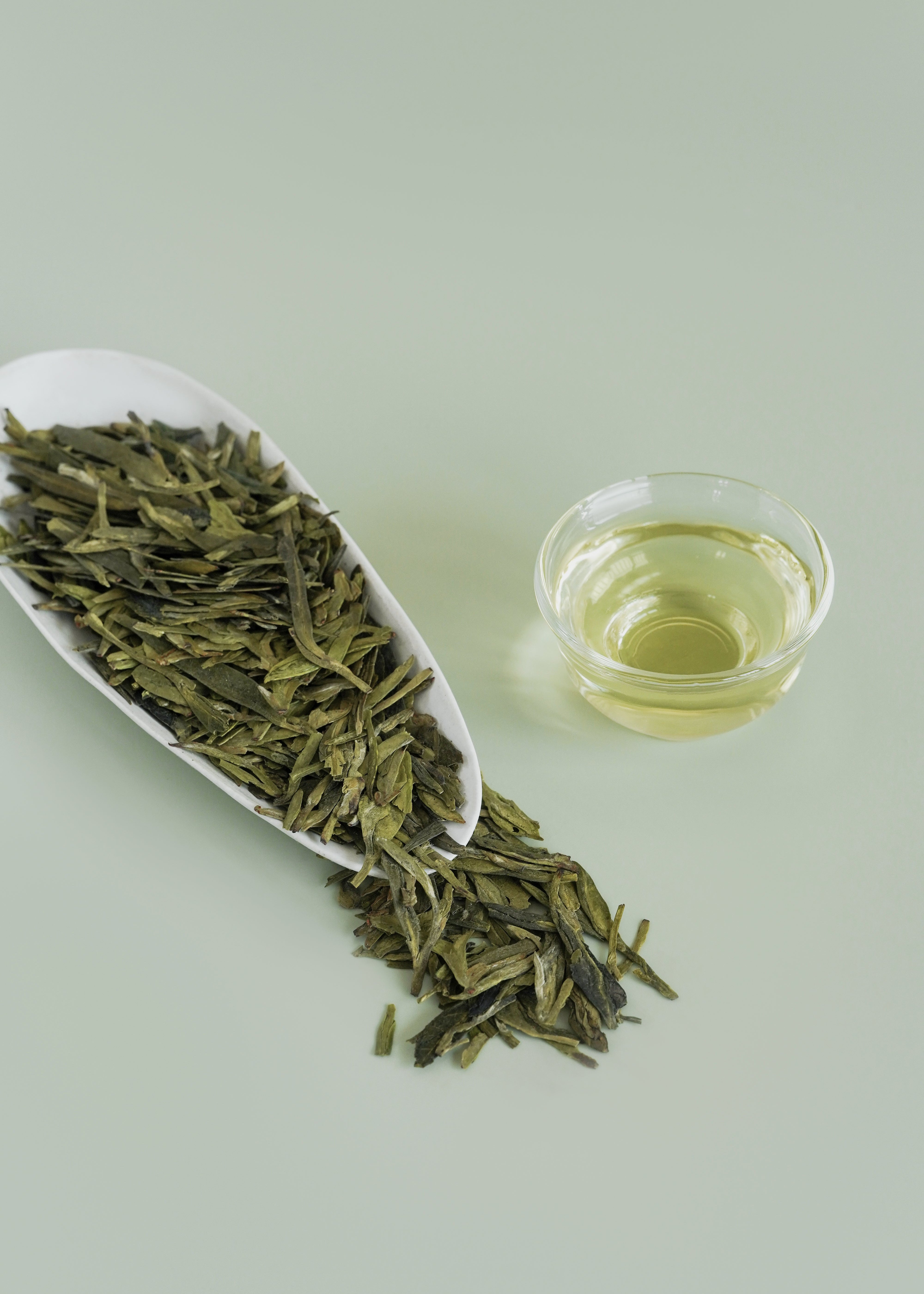 Green tea leaves on a white dish with a small glass of green tea liquid on a light green background. Dragon well longjing