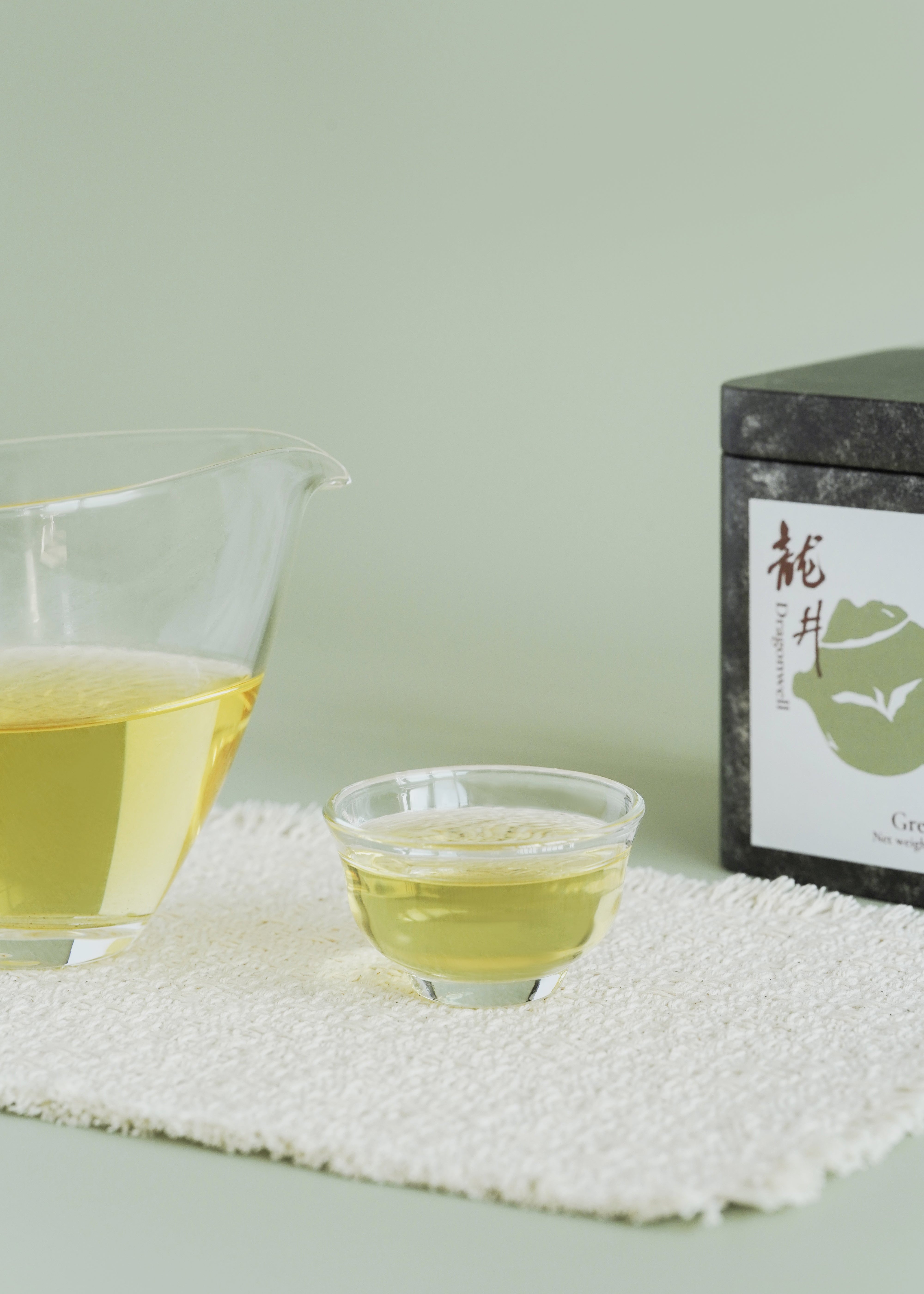 Glass cup with green tea and a box of tea on a textured surface. Dragon well longjing