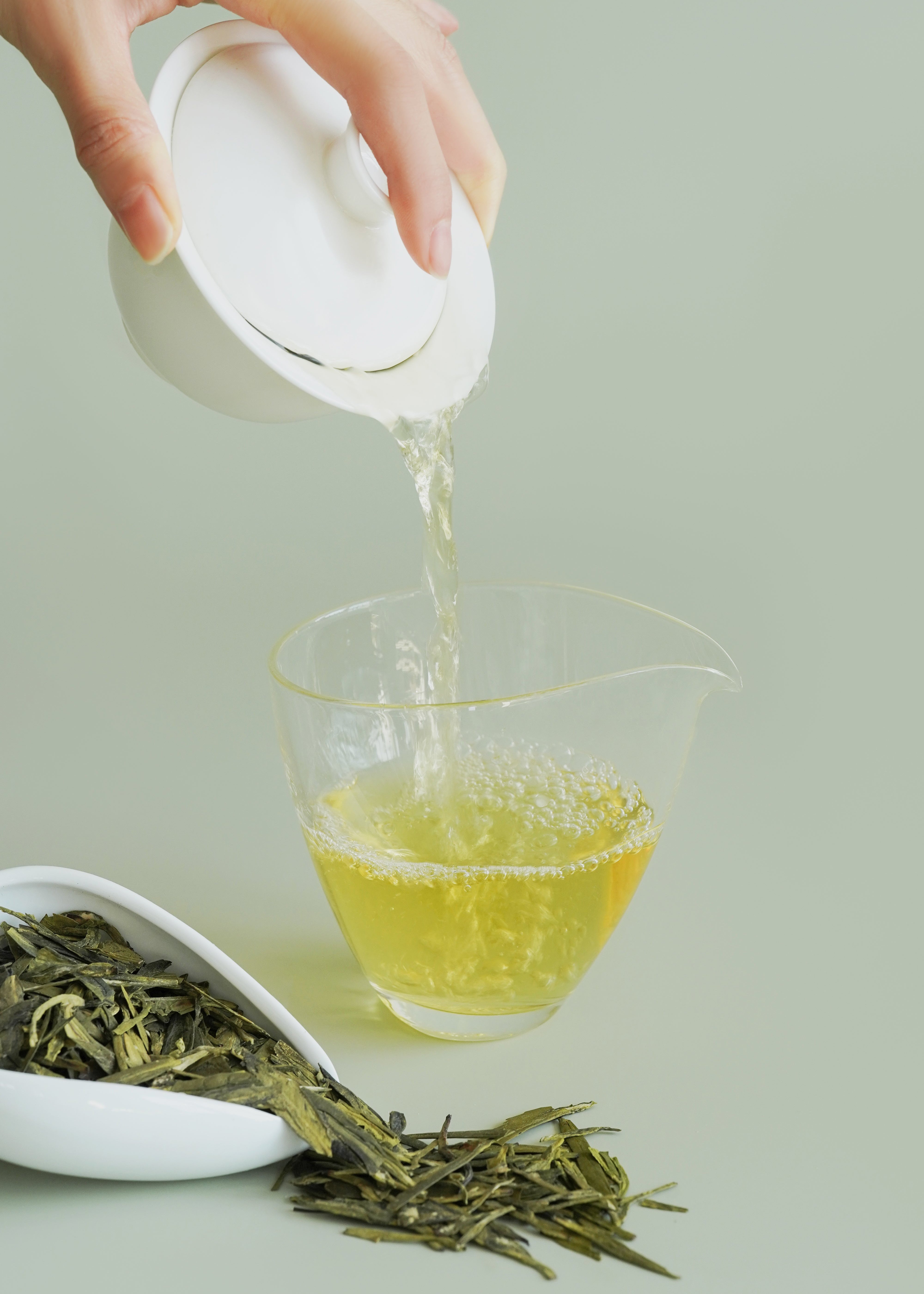 Person pouring green tea into a glass with tea leaves and a white scoop on a light background. Dragon well longjing