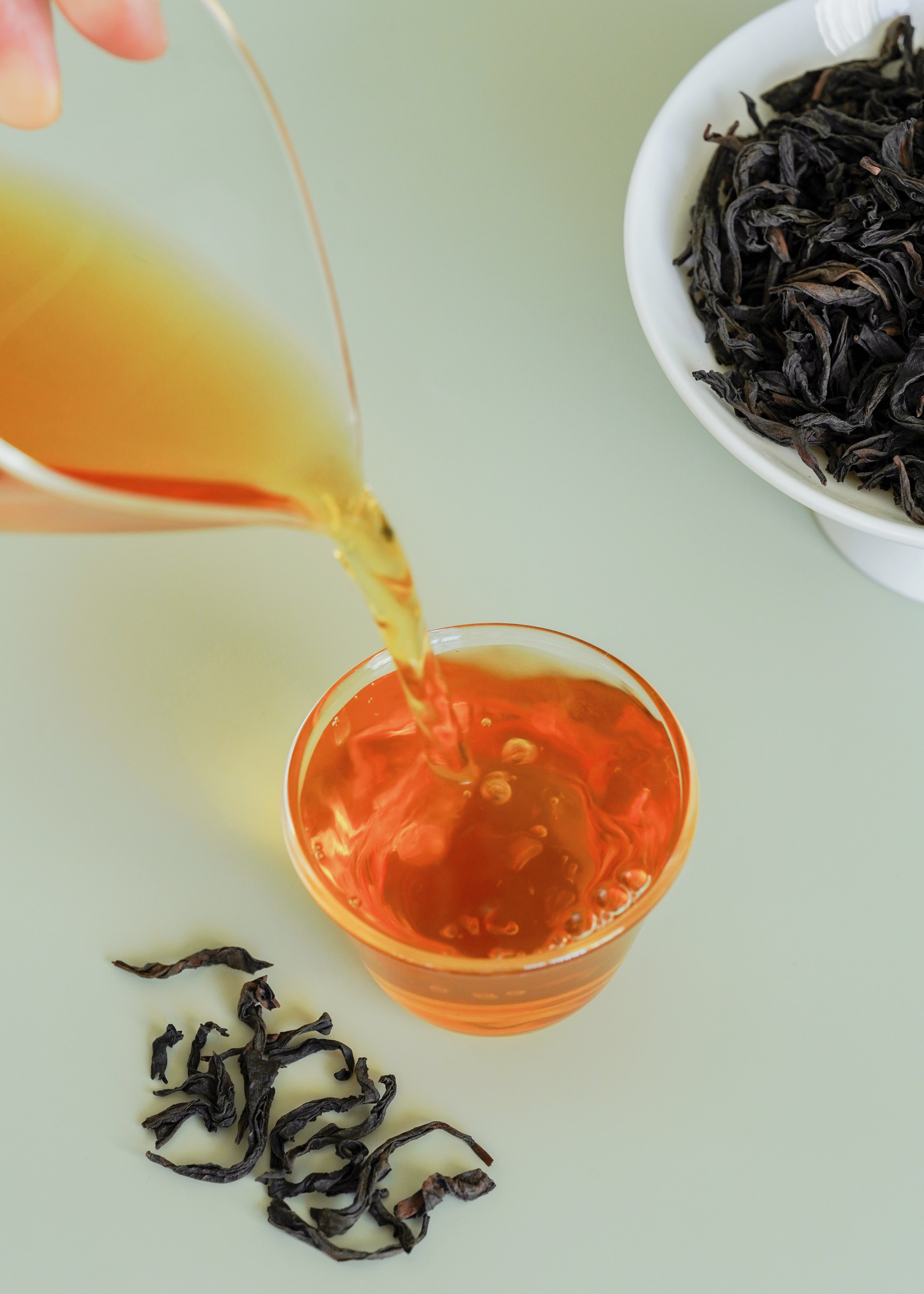 Tea being poured into a glass with tea leaves and a bowl of tea on a light green background. Big red robe, da hong pao