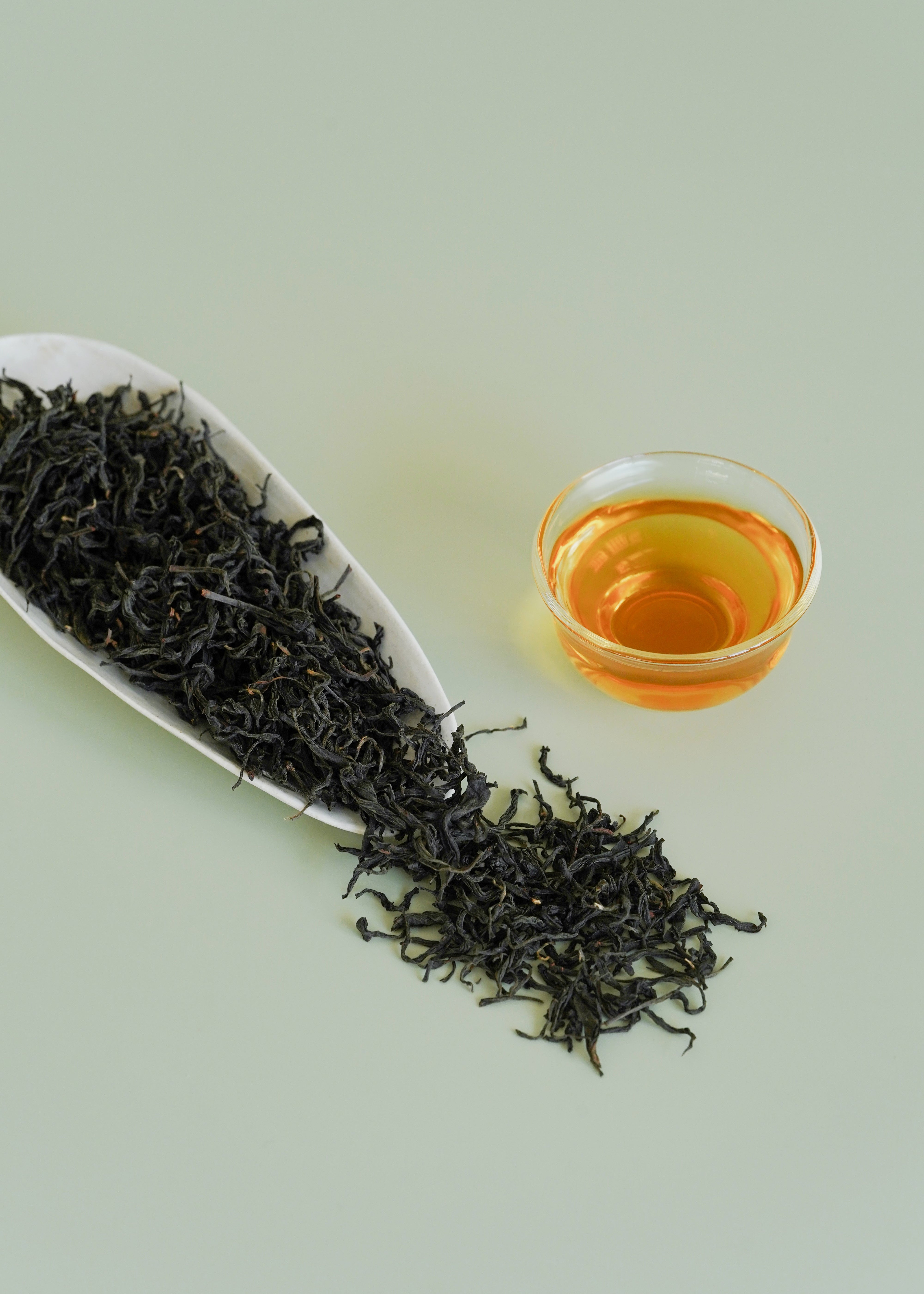 Dried black tea leaves on a white plate with a glass of brewed tea on a light green background. Wild tree black tea