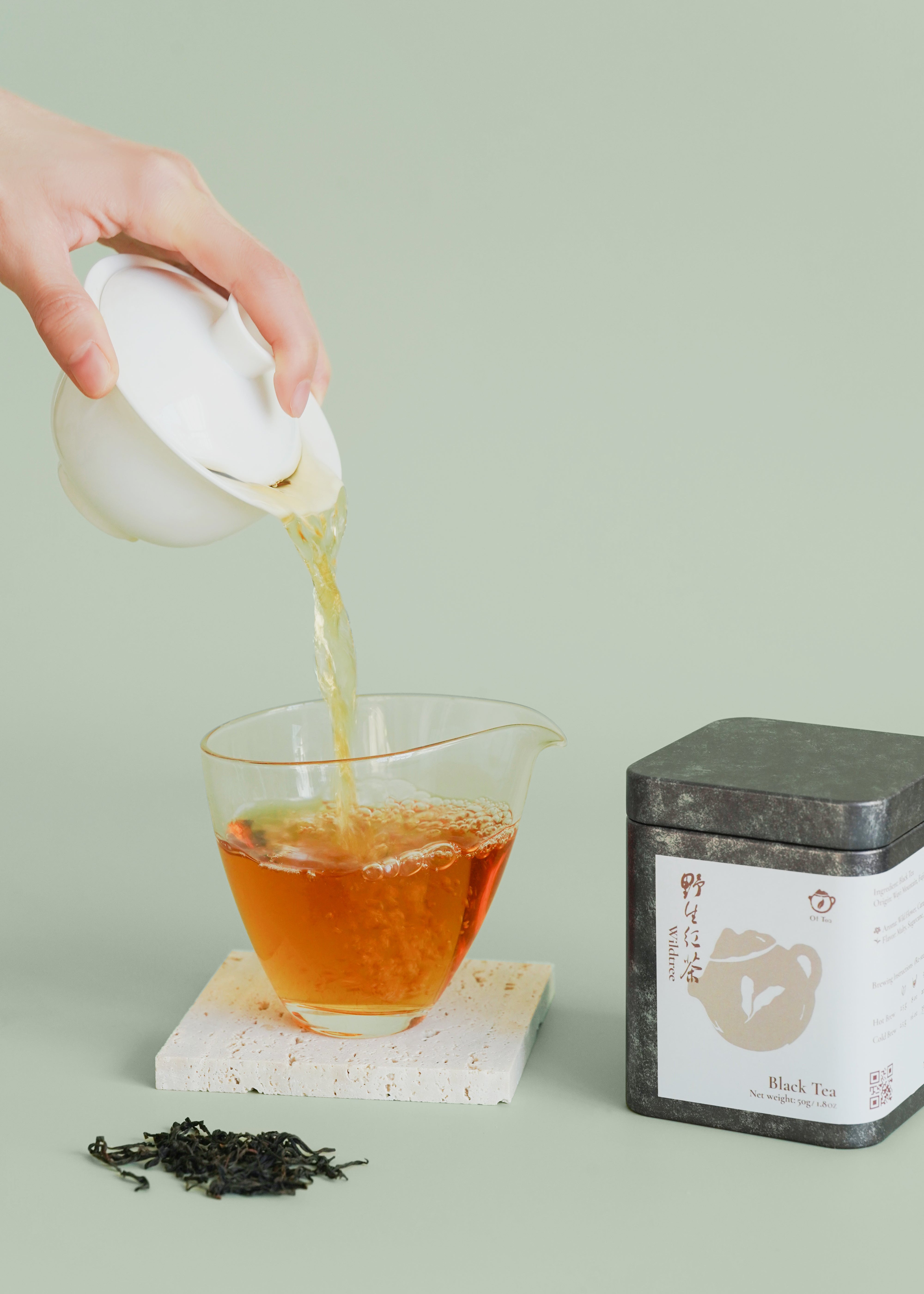 Tea being poured from a white teapot into a glass cup with a tea canister on a light green background. Wild tree black tea