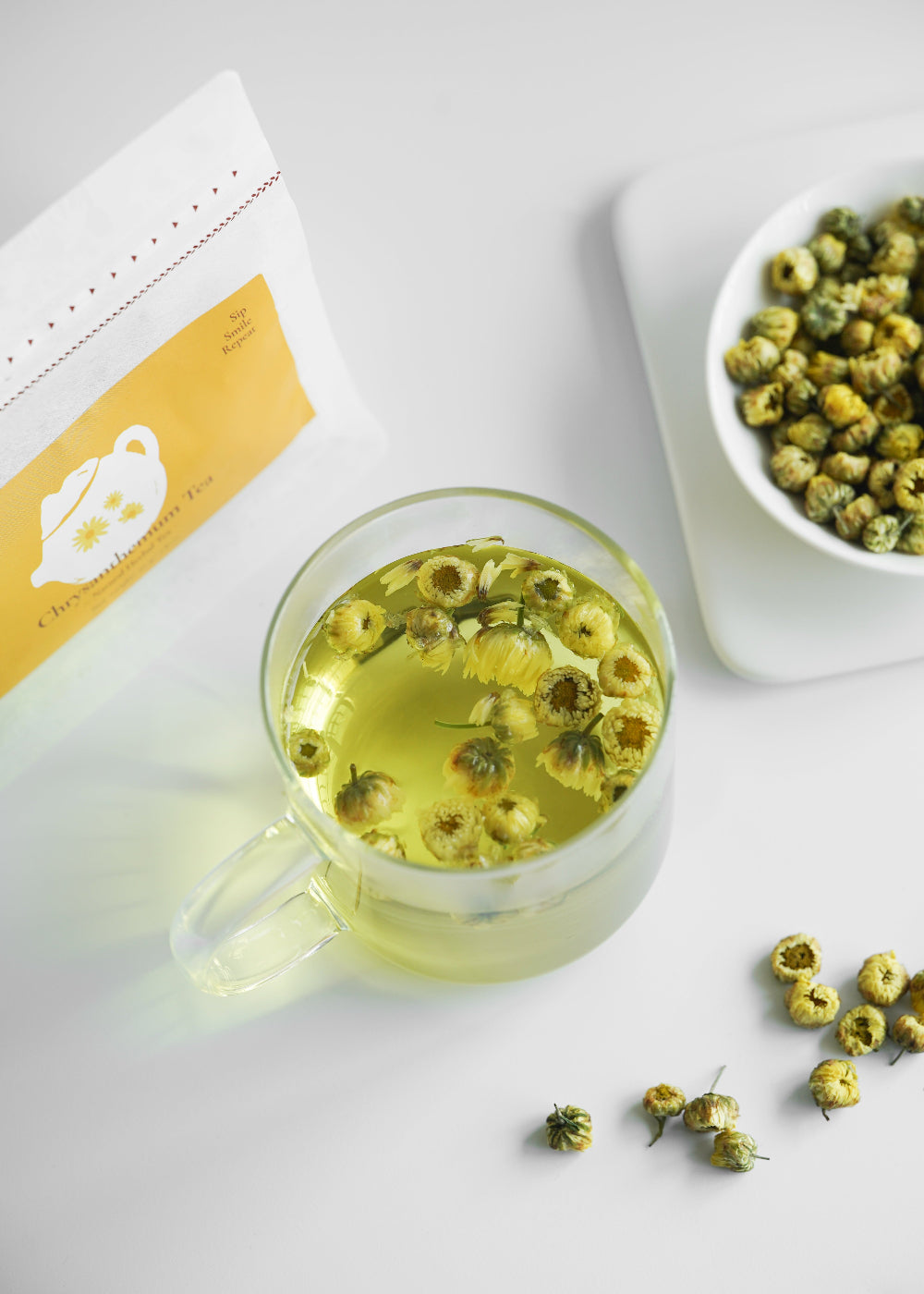 Clear glass mug with yellow floral tea on a white surface, next to a bowl of tea leaves and packaging.