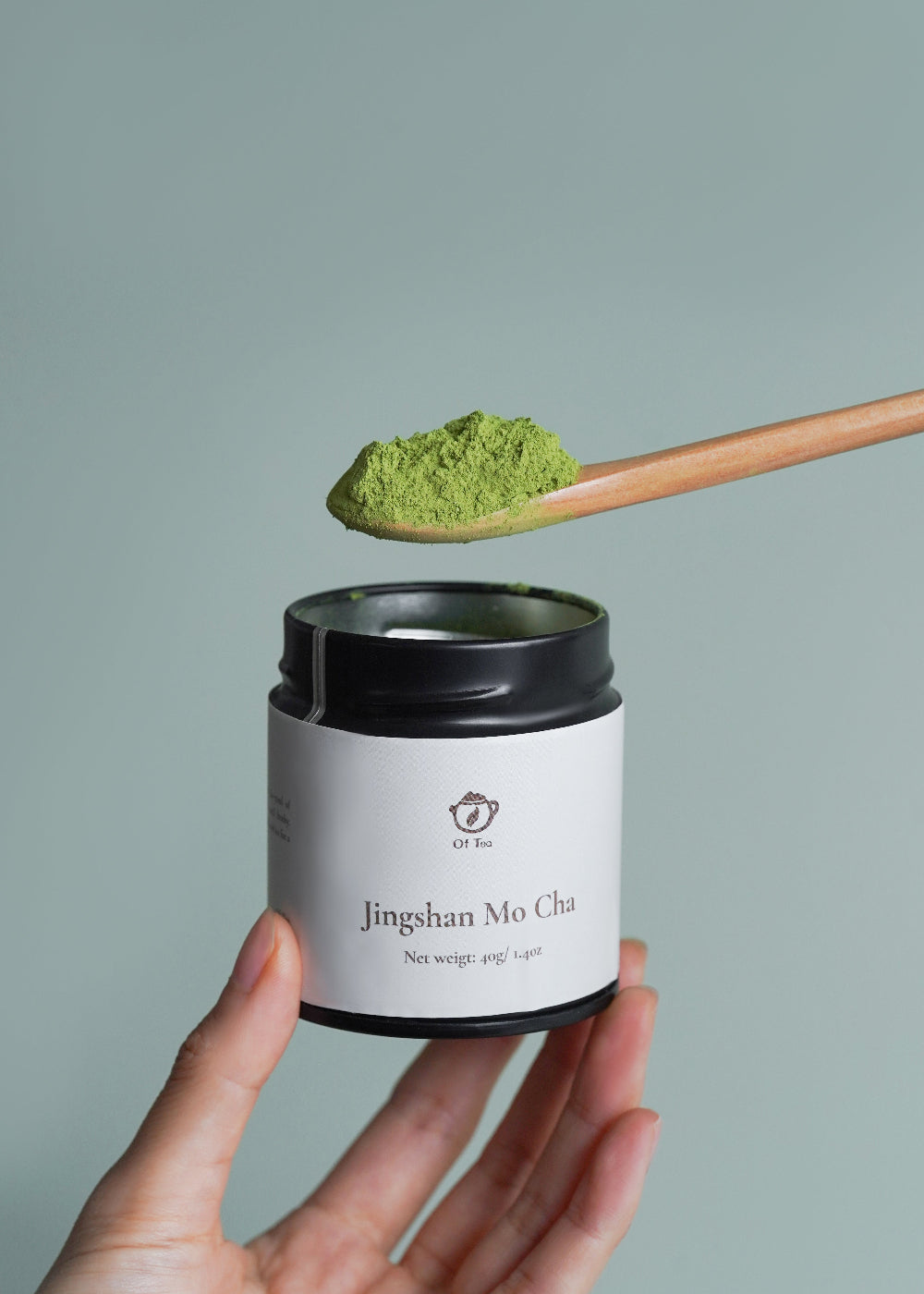 Hand holding a container of Jingshan Mo Cha with a spoonful of green powder above it against a gray background
